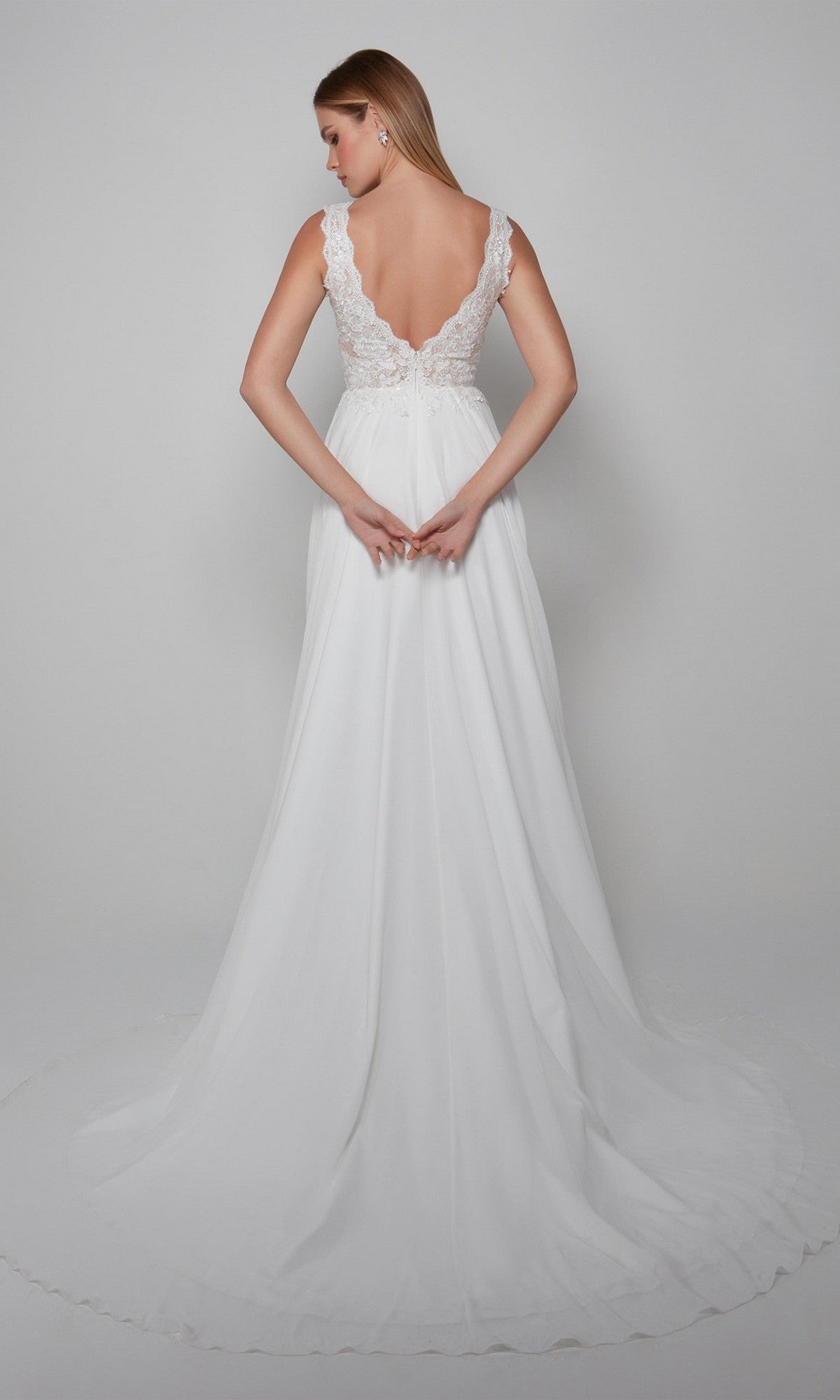 A woman wearing the Alyce 7071 long white dress—a sleeveless lace chiffon gown—stands with her back to the camera, showcasing its deep V-back and flowing train against a plain backdrop.